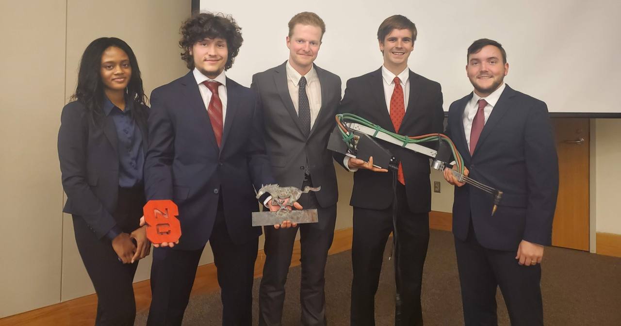 The senior design team at their capstone presentation, holding the first Mach 1 prototype alongside pieces of cut steel