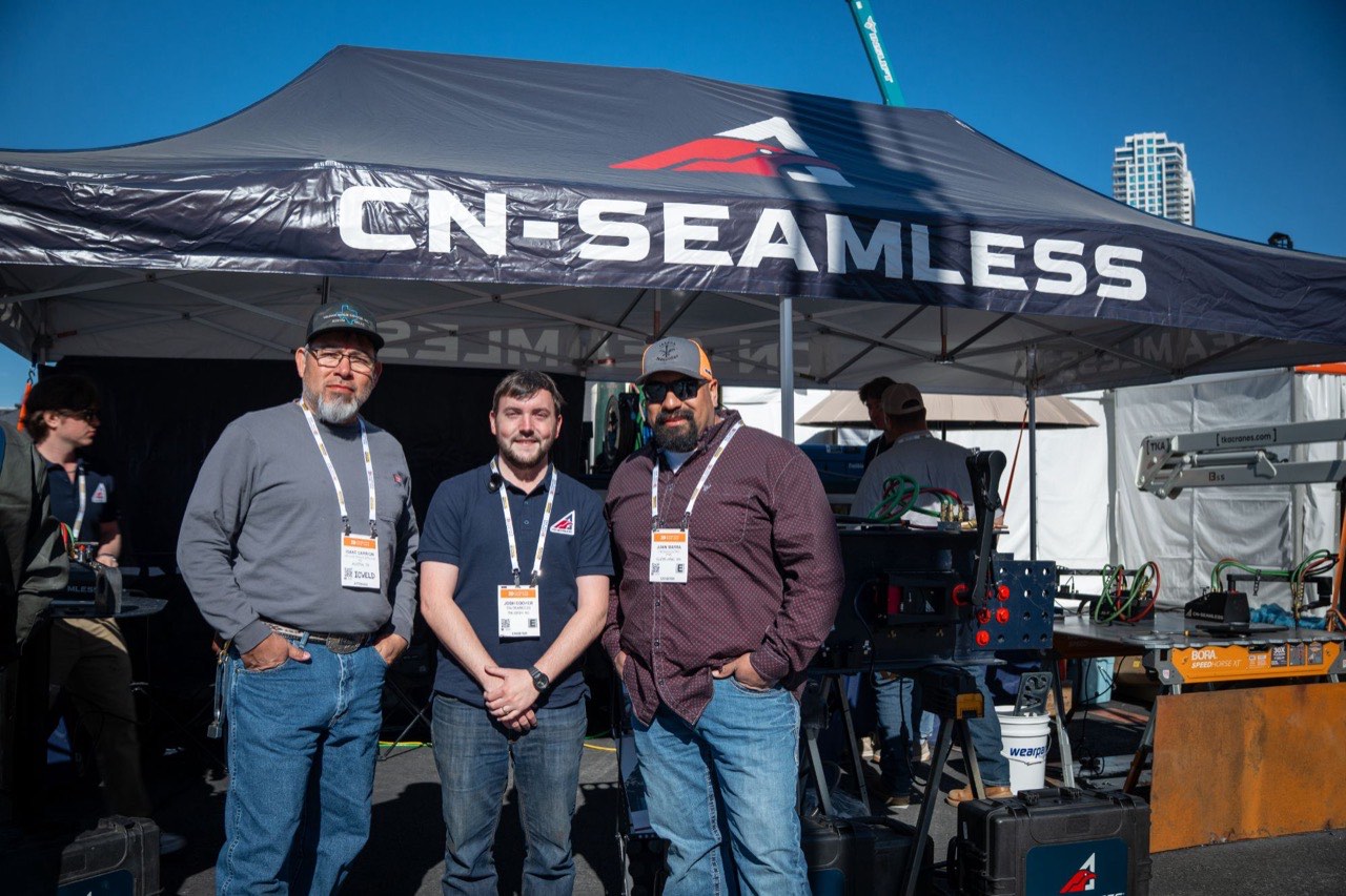 Isaac Carrion, Josh Cooper and Juan Ibarra under the CN-Seamless canopy at Conexpo 2026, with a Mach 1 set up on the bench behind them