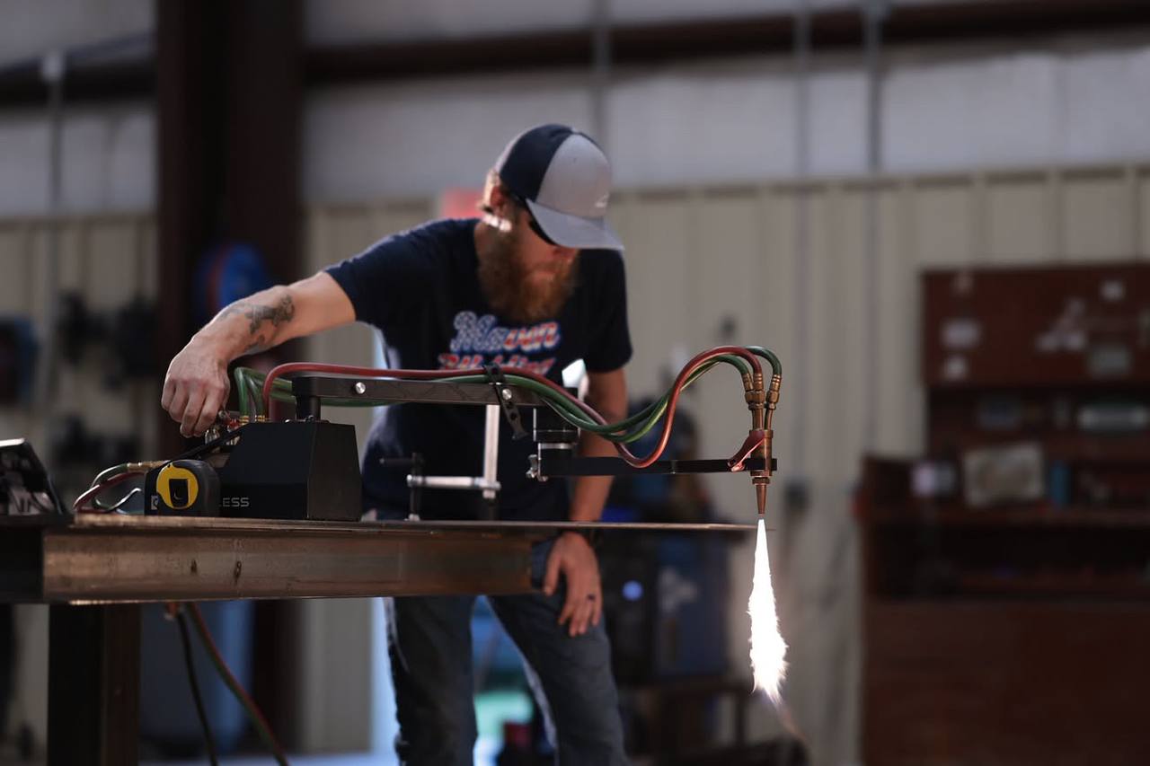 A fabricator steadying a Mach 1 on a steel beam while its oxy-fuel torch cuts, sparks streaming off the underside